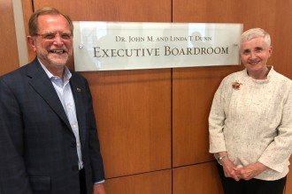Photo of Dr. John and Linda Dunn in front of a sign that reads Dr. John M. and Linda T. Dunn Executive Board Room.
