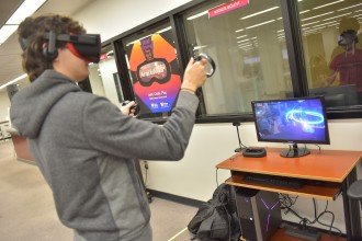 A student wears virtual reality goggles and holds up two controllers while watching a nearby computer screen.