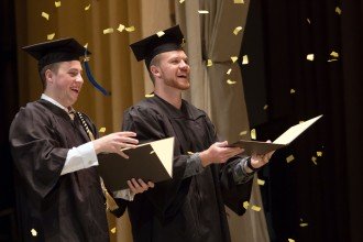 Two male students in graduation caps and gowns use their diploma folders to catch some of the confetti being dropped during their commencement ceremony.