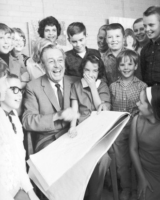A black-and-white photo of a seated Walt Disney surrounded by children. On his lap, Disney is holding a large tablet of paper.