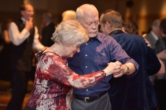 A senior couple (male and female) lock arms and dance together on a crowded dance floor.