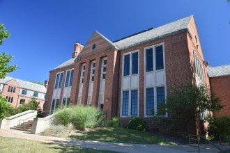 This is a photo of the exterior of Walwood Hall. The building is two stores tall on the west side, and three stories tall on the east side. The sides of the building are red brick and they are topped with a grey roof. Wallwood hall was built in 1939 for the dual role as a student union and the first dormitory on campus.