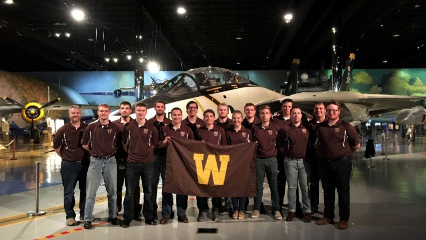 Members of the 2017 regional Sky Broncos team standing in a hangar in front of an airplane holding a W flag.