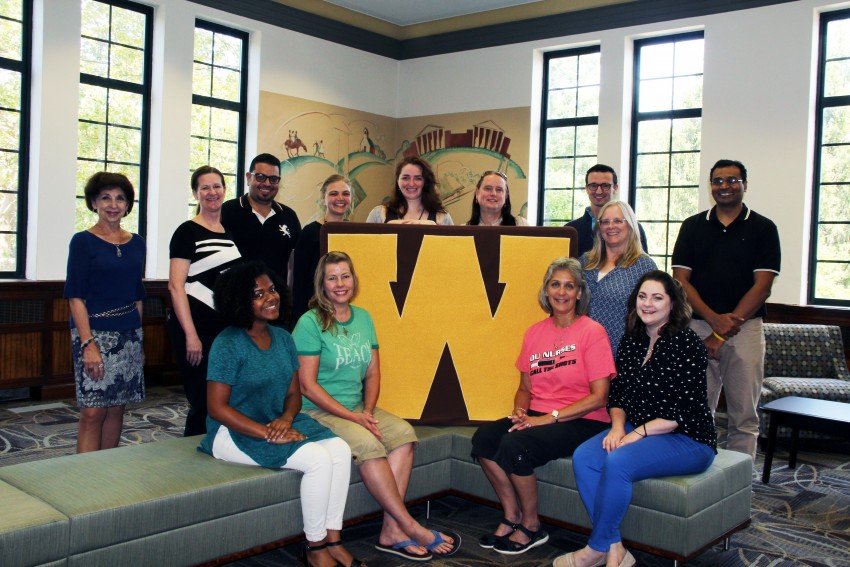 2017 Dissertation Cafe participants with Dr. Christine Byrd-Jacobs, Acting Dean of the Graduate College and Dr. Di Pierro, Program Manager for Graduate Research and Retention. A group of eleven diverse students surround a large gold W which pronounces the WMU brand. Walwood Hall, where the Graduate College is located, has been recently renovated with new carpet and furniture to host small and large groups. The original murals from 1939 can be seen behind the students, while the tall narrow windows frame a view of East Campus with its many trees and classic buildings.