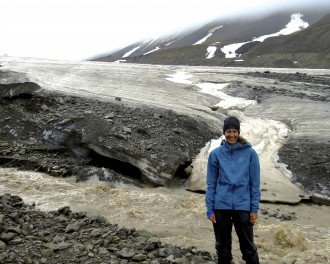 Photo of McNeal standing on the rock-strewn fringe of a melting glacier with a stream of rushing water swirling at her feet.