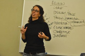 a photo of a professor at the front of a whiteboard in a classroom with a marker in one hand and an eraser in the other. She is explaining the list of terms that she has written on the board.